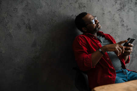 Black man in eyeglasses using cellphone while working at desk indoors