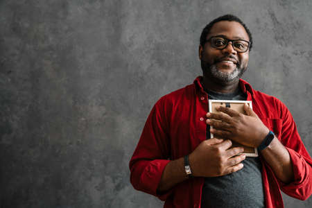 Black man in eyeglasses smiling and holding photo frame while standing by concrete wall indoorsの写真素材