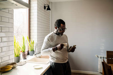 Black man wearing eyeglasses using mobile phone while drinking coffee at homeの写真素材