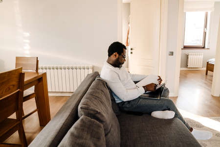 Black man wearing eyeglasses working with laptop and papers on sofa at homeの写真素材