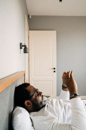 Black man wearing eyeglasses using mobile phone while resting in bed at homeの写真素材