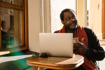 Black bearded man using mobile phone and laptop while sitting in cafe outdoorsの写真素材