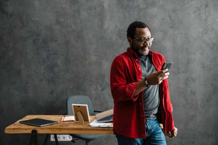 Black man in eyeglasses using cellphone while standing by desk indoorsの写真素材