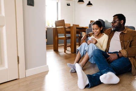 Black girl and her father hugging and using cellphone while sitting on floor at homeの写真素材
