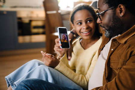 Black girl and her father hugging and using cellphone while sitting on floor at homeの写真素材