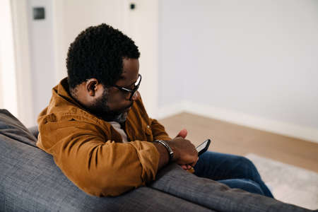 Black bearded man using cellphone while sitting on sofa at homeの写真素材