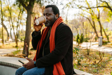 Black man using cellphone and drinking coffee while sitting on bench in parkの写真素材