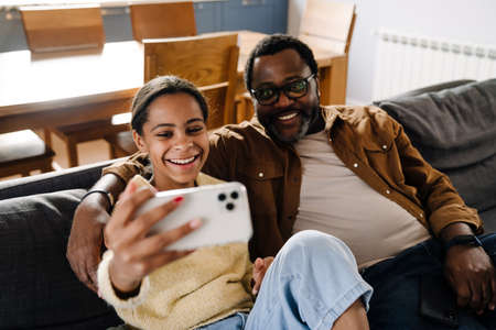 Black girl and her father taking selfie on cellphone while sitting on sofa at homeの写真素材