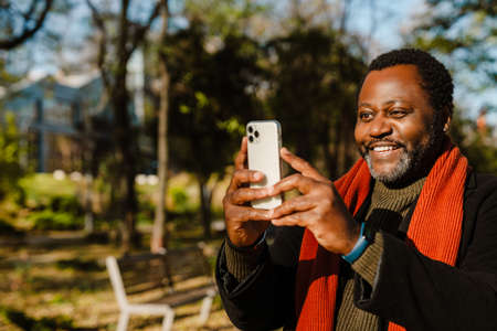 Black bearded man taking photo on cellphone while strolling in park outdoors