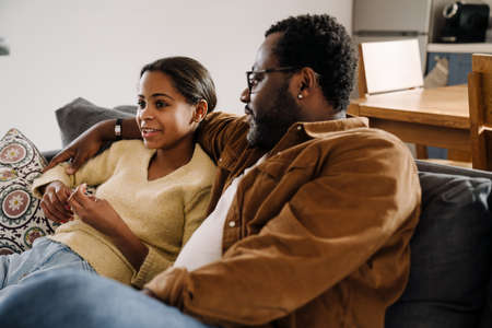 Black girl talking and smiling with her father while sitting on sofa at homeの写真素材