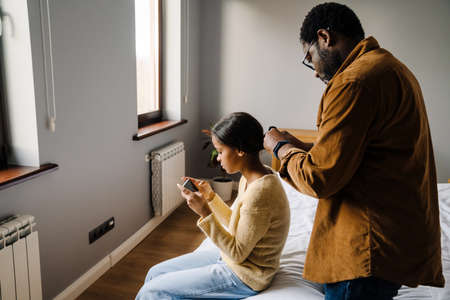 Black girl using cellphone while father doing her hairstyle at homeの写真素材