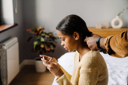 Black girl using cellphone while father doing her hairstyle at homeの写真素材