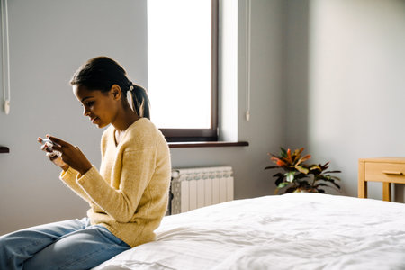 Black brunette girl using mobile phone while sitting on bed at homeの写真素材