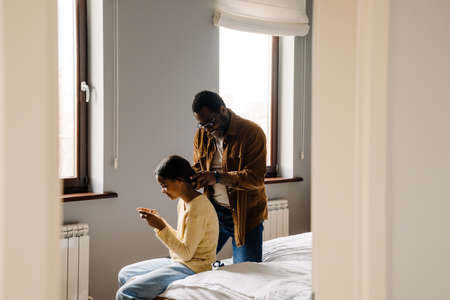 Black girl using cellphone while father doing her hairstyle at homeの写真素材