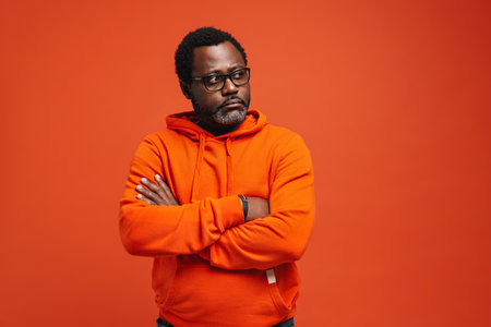 Black sad man in eyeglasses posing with arms crossed isolated over orange backgroundの写真素材