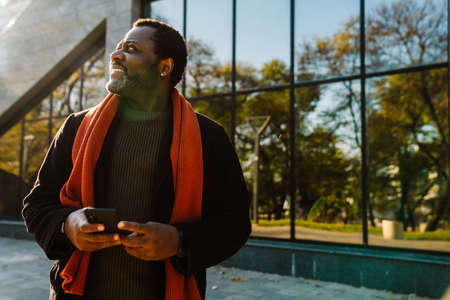 Black bearded man smiling and using cellphone on city streetの写真素材