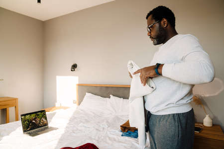 Black man using laptop while folding his clothes in bedroom at homeの写真素材