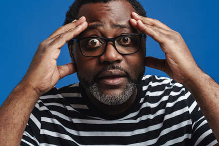 Black bearded man in eyeglasses expressing surprise at camera isolated over blue backgroundの写真素材