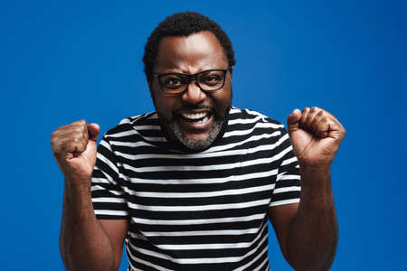 Black man in eyeglasses screaming while making winner gesture isolated over blue backgroundの写真素材