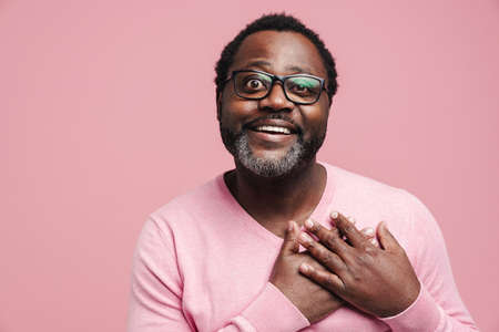 Black man in eyeglasses smiling while holding his hands on chest isolated over pink backgroundの写真素材
