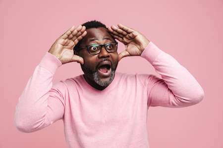 Black man in eyeglasses screaming while expressing surprise at camera isolated over pink backgroundの写真素材