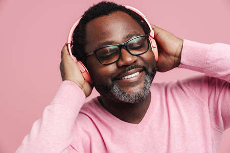 Black bearded man smiling while listening music with headphones isolated over pink backgroundの写真素材