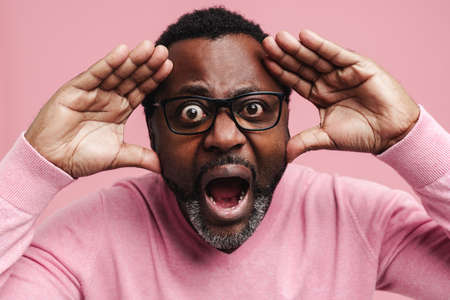 Black man in eyeglasses screaming while expressing surprise at camera isolated over pink backgroundの写真素材