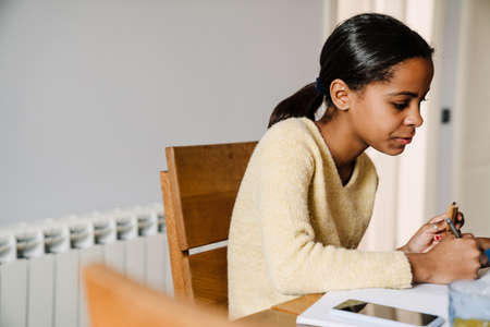 Black happy girl eating cookie while doing homework in kitchen at homeの写真素材
