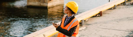 Black woman wearing helmet and vest using mobile phone while working in portの写真素材