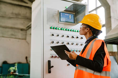 Black woman wearing helmet and vest working with equipment at factoryの写真素材