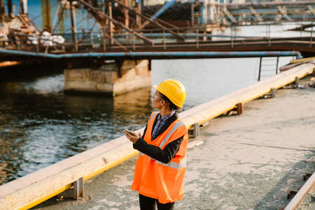 Black woman wearing helmet and vest using mobile phone while working in portの写真素材
