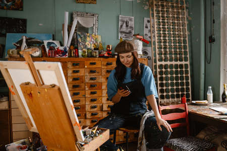 Young woman wearing overall using mobile phone while working in her studio