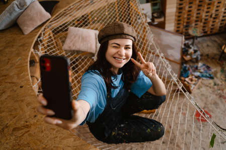 Young woman artist gesturing while taking selfie on cellphone in her studio indoors