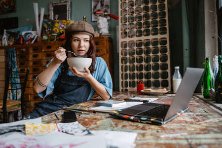 Young woman artist eating cereal while working with laptop in her studio indoors