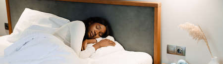 Black young woman sleeping while lying on white bed linen at home