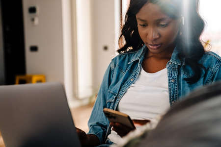 Black young woman in earphones using mobile phone while sitting on couch at home
