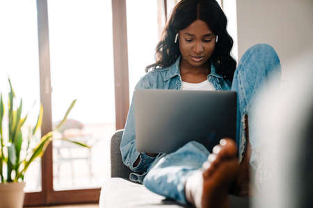 Black young woman in earphones using laptop while sitting on couch at home