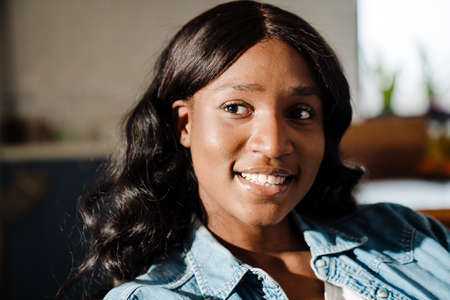 Black young woman wearing denim shirt smiling while sitting on couch at home