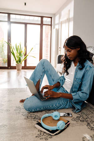 Black young woman in earphones using laptop while sitting on floor at home