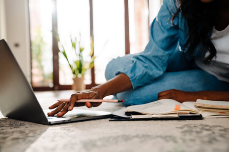 Black young woman studying and using laptop while sitting on floor at homeの写真素材