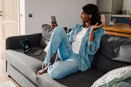 Black young woman in earphones using mobile phone while sitting on couch at homeの写真素材