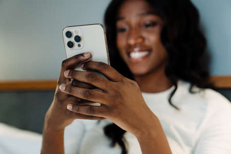 Black young woman smiling and using mobile phone while lying in bed at homeの写真素材