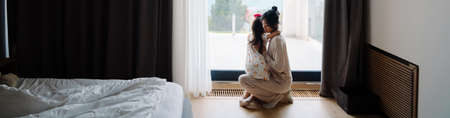 Asian woman and her daughter looking out window together at home