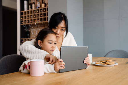 Asian woman and her daughter using tablet computer while sitting at table in homeの写真素材