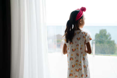 Asian girl wearing home clothes looking out window in bedroom at home