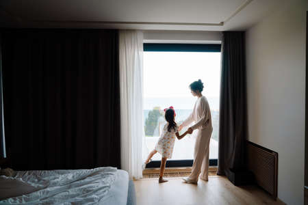 Asian woman and her daughter looking out window together at homeの写真素材