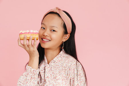 Asian girl wearing headband smiling while posing with doughnut isolated over pink backgroundの写真素材