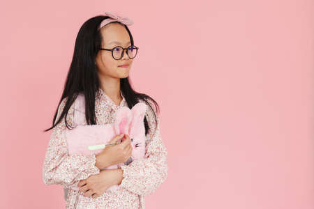 Asian girl wearing eyeglasses posing with stuffed exercise book isolated over pink background