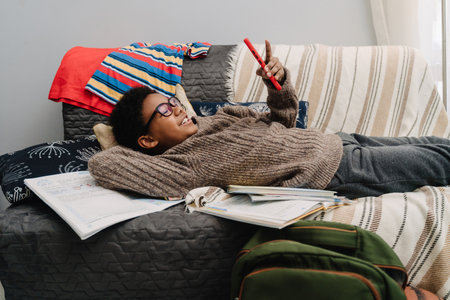 Black boy in eyeglasses laughing and using cellphone while resting at homeの写真素材