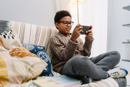 Black boy in eyeglasses playing online game on cellphone while resting at homeの写真素材